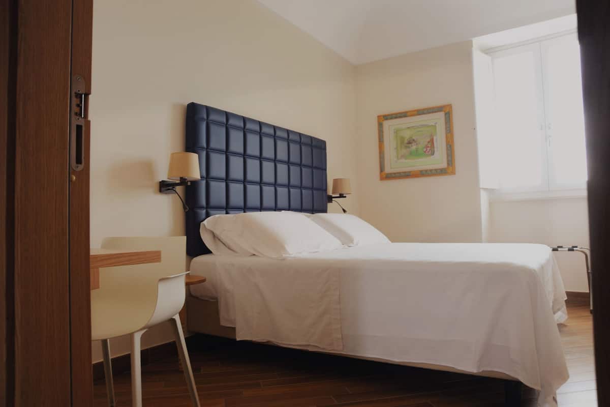 Un dormitorio con cama, lámparas de noche, una silla y una ventana.