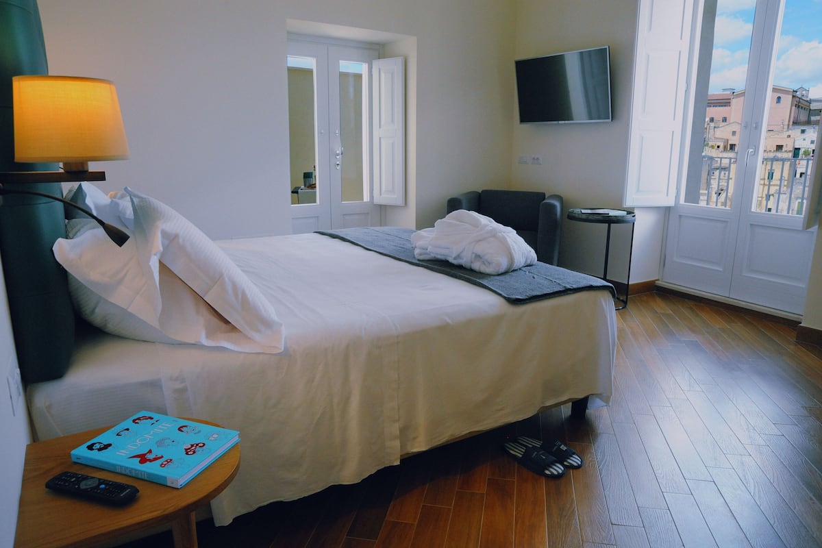 Habitación de hotel con cama, televisor, silla y una mesita con un libro y control remoto.