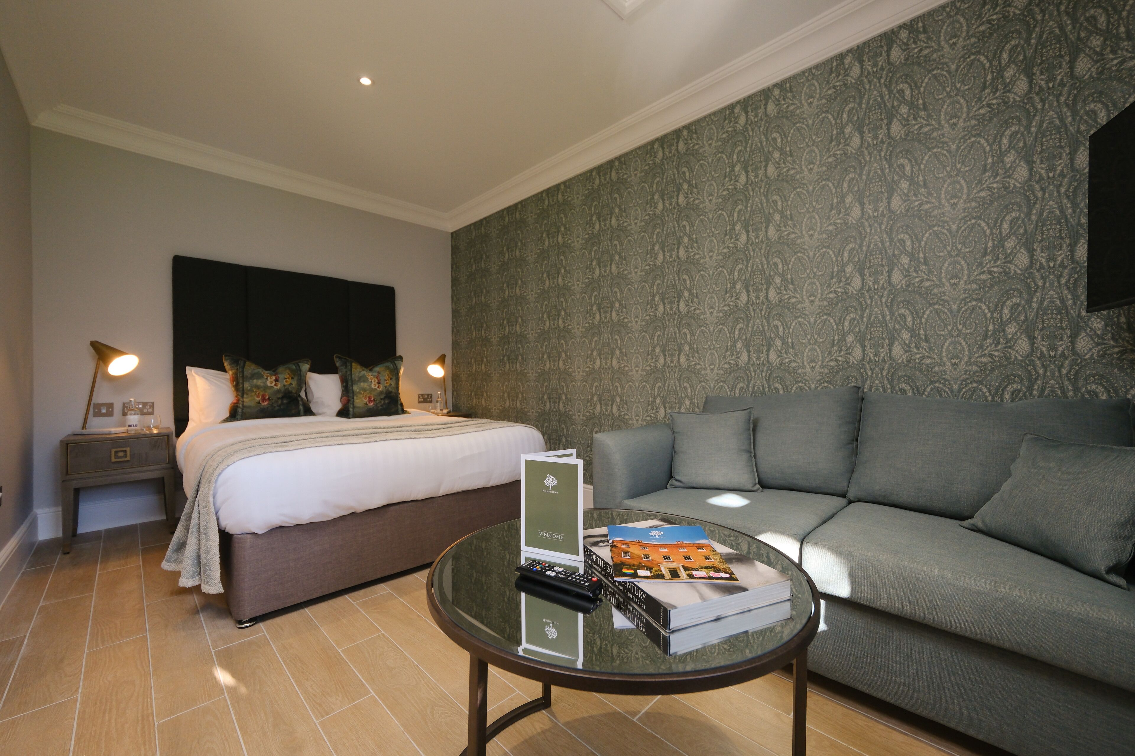 Une chambre d’hôtel comprenant un lit, un canapé, une table basse en verre avec une télécommande et des livres, ainsi qu’une table de chevet avec une lampe.