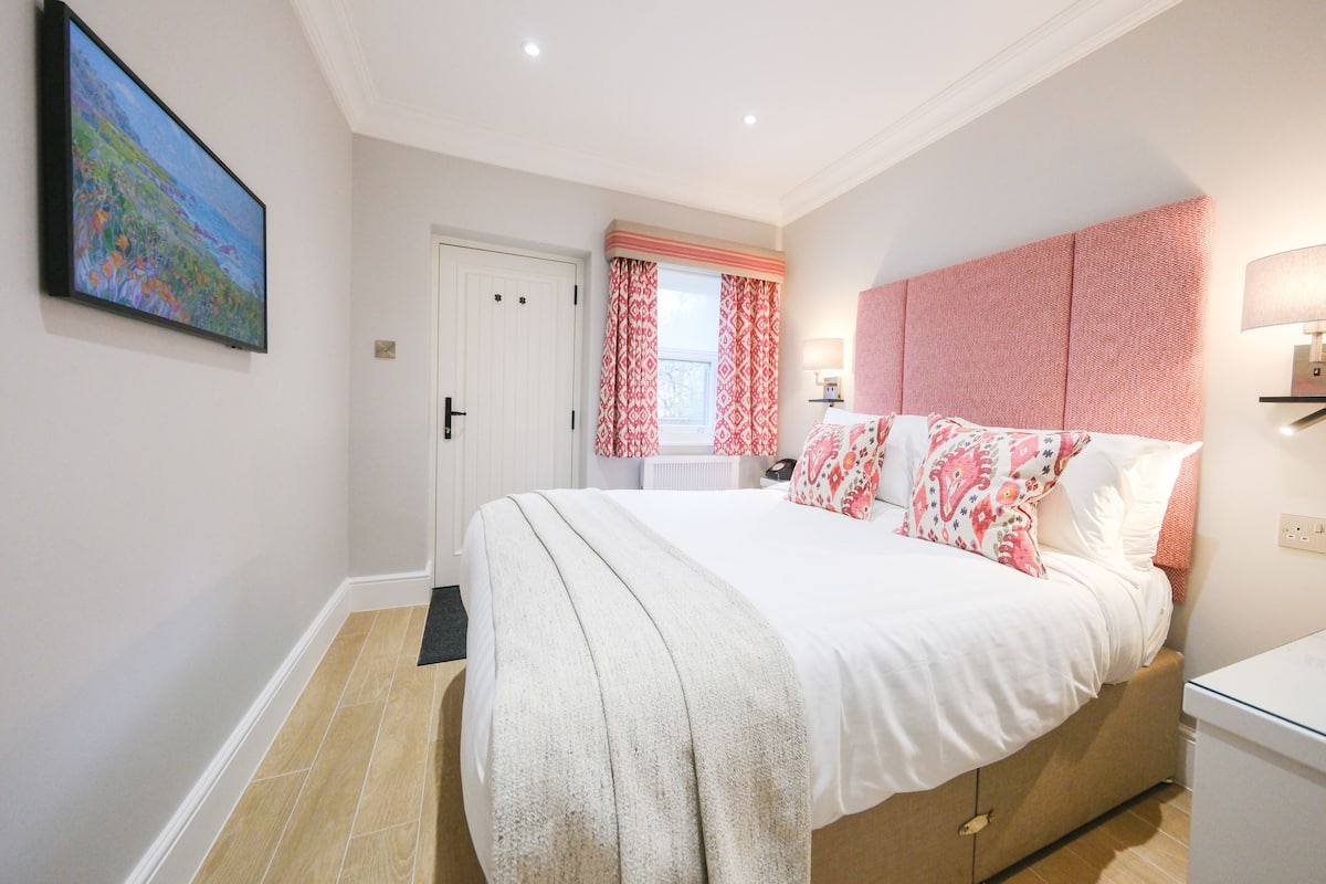 A bedroom with a bed, a bedside table, a lamp, a window with curtains, and a painting on the wall.