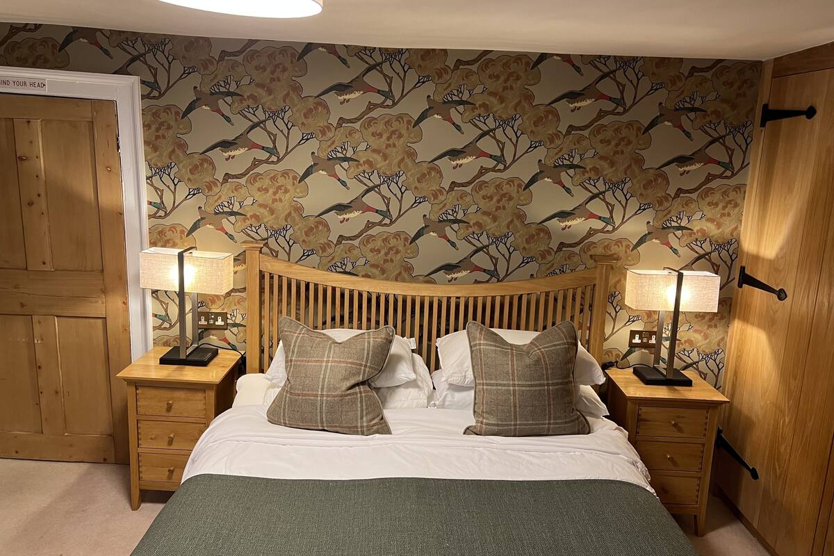 A bedroom with a wooden headboard, two bedside tables with lamps, a bed with white linens and plaid pillows, and a floral wallpaper.