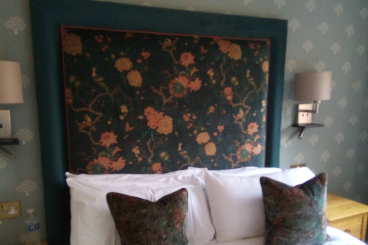 A bedroom with a floral headboard, two pillows, a bedside table, and a lamp.