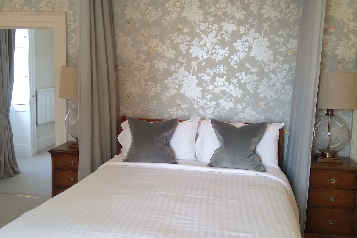 A neatly made bed with a tufted headboard, a wooden nightstand with a lamp, and a floral-patterned wallpaper.