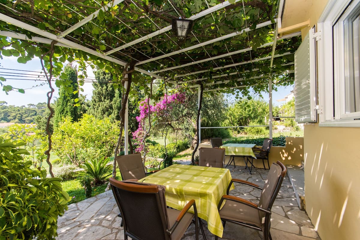 Een terras buiten met een tafel en stoelen, omgeven door groen en een pergola.