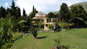 Une maison à deux étages dotée d’un balcon, entourée d’une végétation luxuriante et d’une cour verdoyante.