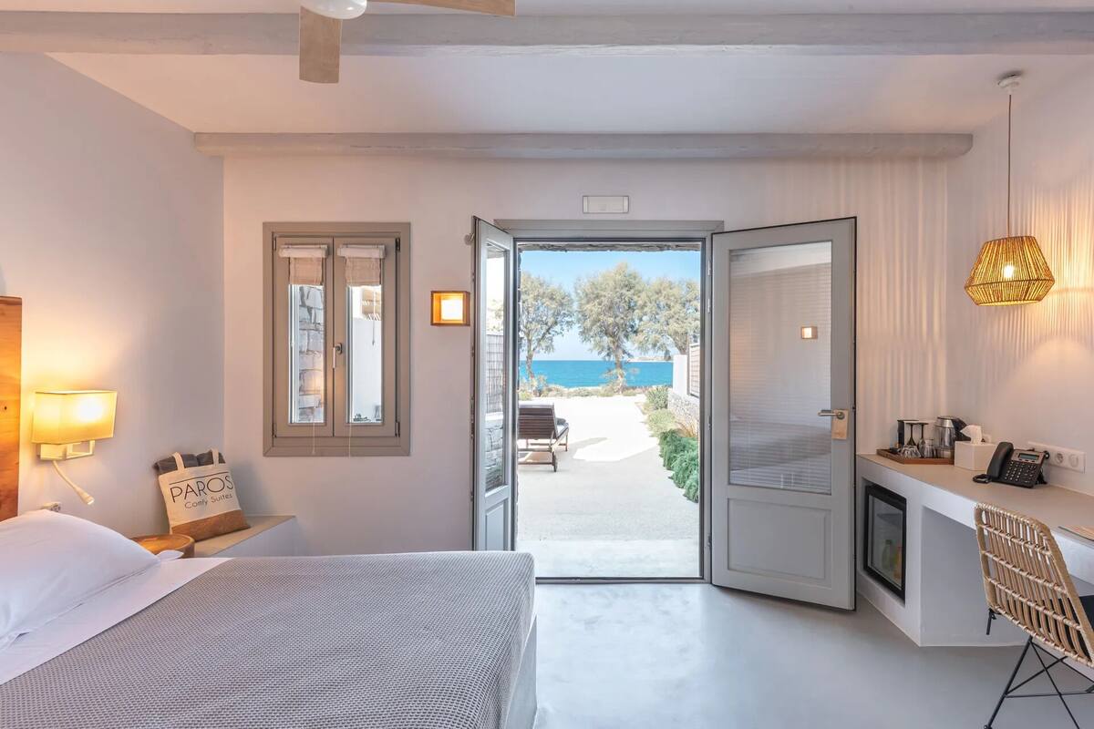 Ein Schlafzimmer mit Bett, Schreibtisch, Stuhl, Lampe und Meerblick durch eine offene Tür.