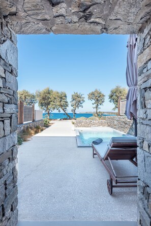 A stone archway leading to a pool area with a view of the sea, surrounded by trees and stone walls. - Paros Comfy Suites (Paros)