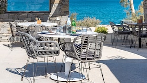Un patio al aire libre con paredes de piedra, sillas de mimbre y una mesa lista para comer. Un ventanal grande ofrece vistas al mar.