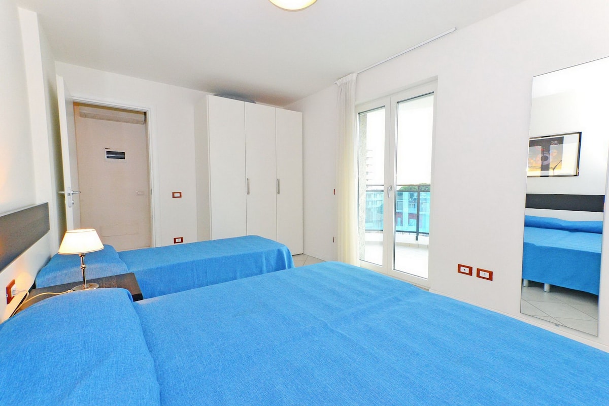 A hotel room with two beds, a nightstand, a lamp, a sliding door to a balcony, and a large mirror.