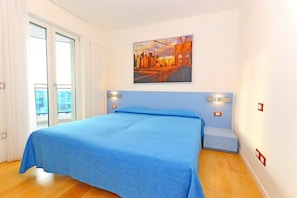 A bedroom with a large bed, a cityscape painting on the wall, and a view through a sliding door. - Residence Sky (Lignano Sabbiadoro)