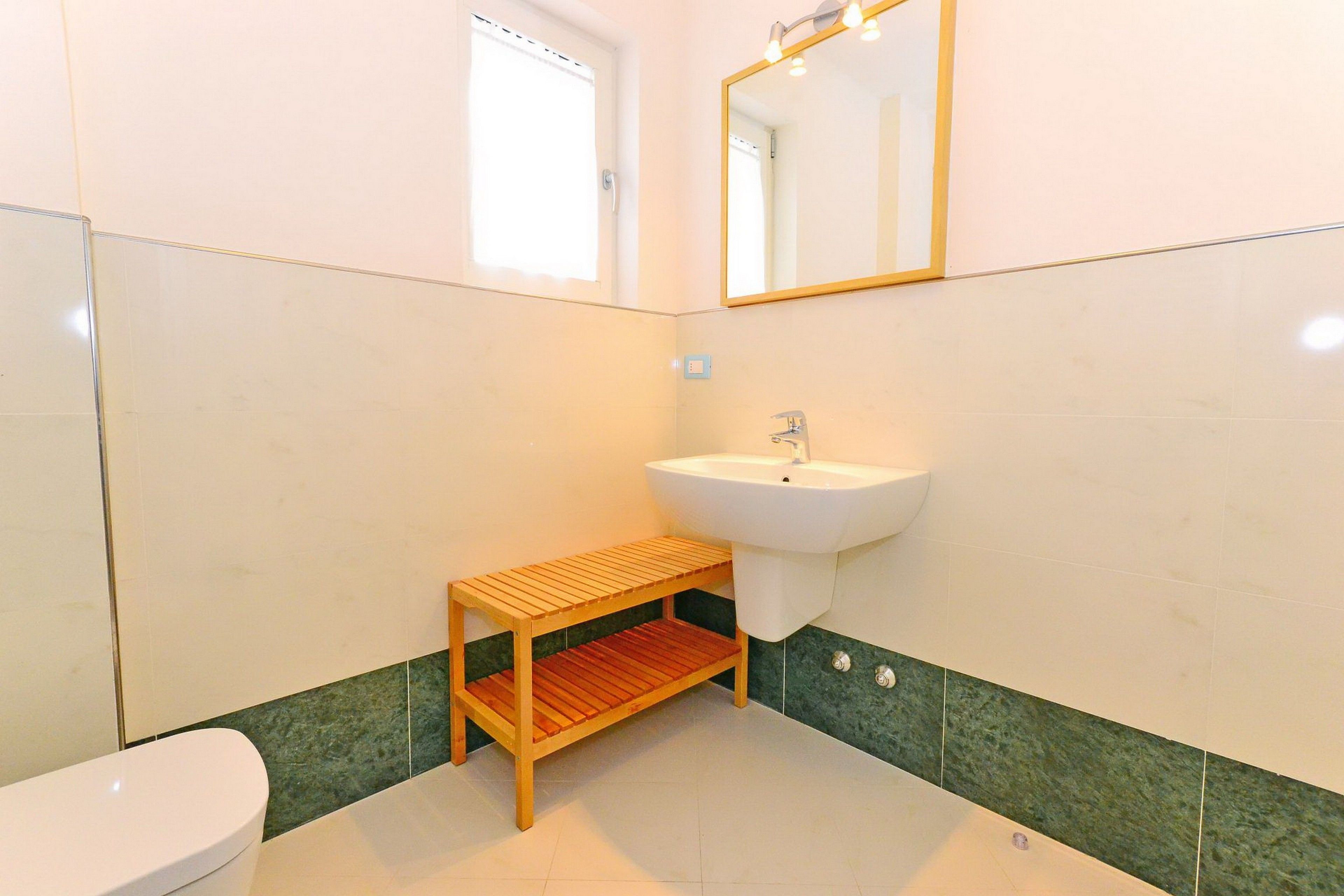Un baño con inodoro, lavabo, estante de madera, espejo y paredes con azulejos verdes.