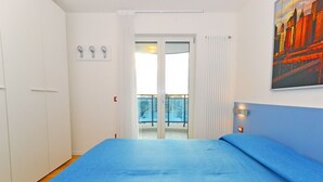 A bedroom with a large bed, a balcony with a view, white walls, and a cityscape painting. - Residence Sky (Lignano Sabbiadoro)
