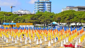 A beach with rows of beach chairs and umbrellas, a building with balconies, and trees in the background. - Residence Sky (Lignano Sabbiadoro)