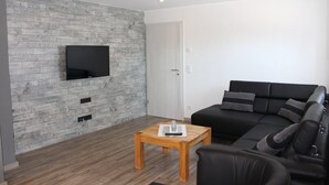 A modern living room with a black leather sofa, a wooden coffee table, and a flat-screen TV mounted on a brick-patterned wall.