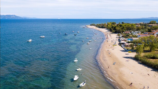 Una playa costera con barcos fondeados cerca de la orilla, arena y una hilera de edificios con techos rojos.
