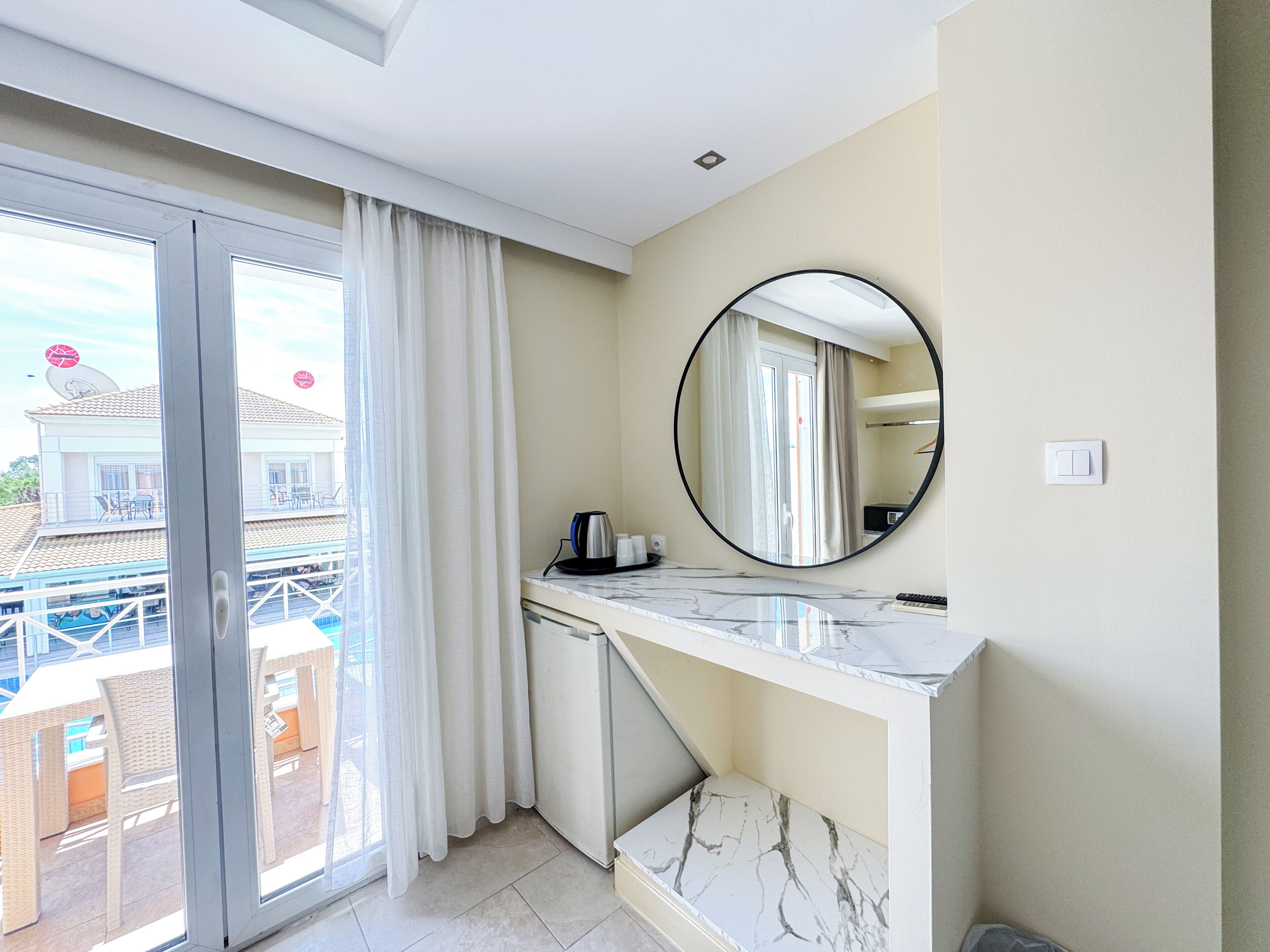 Quarto de hotel com porta de vidro deslizante para uma varanda, um espelho redondo na parede e bancada de mármore.
