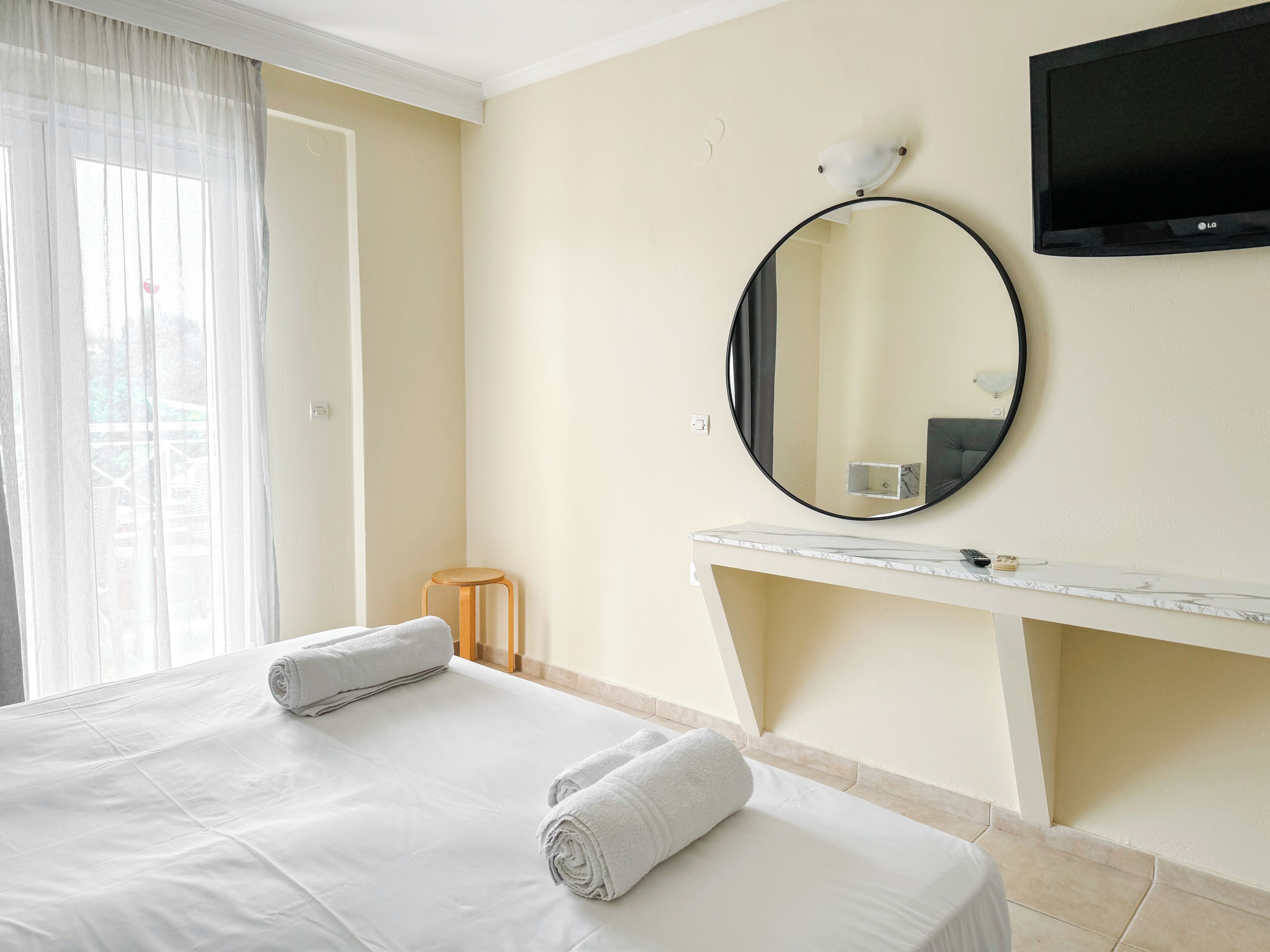 A hotel room with a bed, a round mirror, a TV, and a small stool.