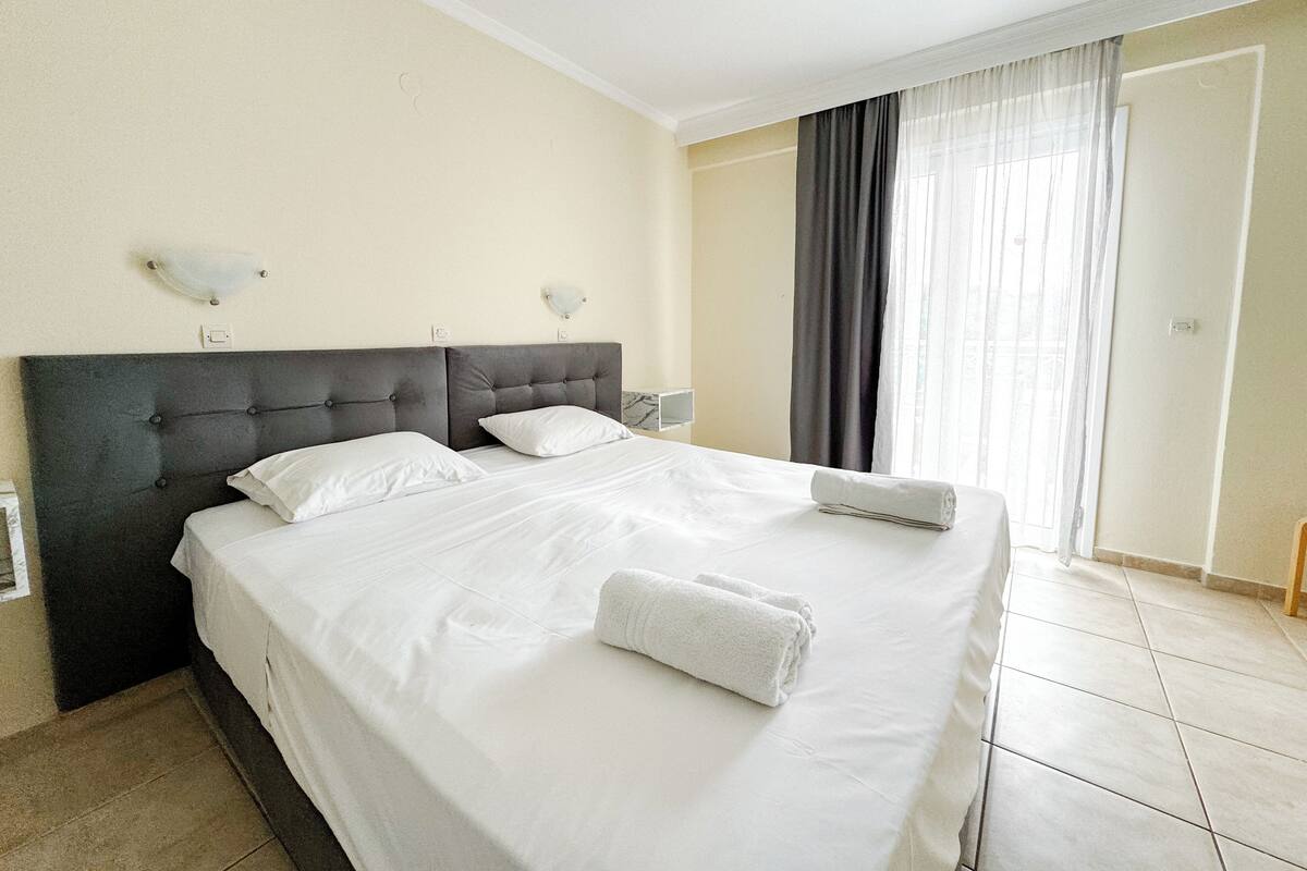A neatly made bed with white linens and a folded towel, a dark headboard, and a window with curtains.