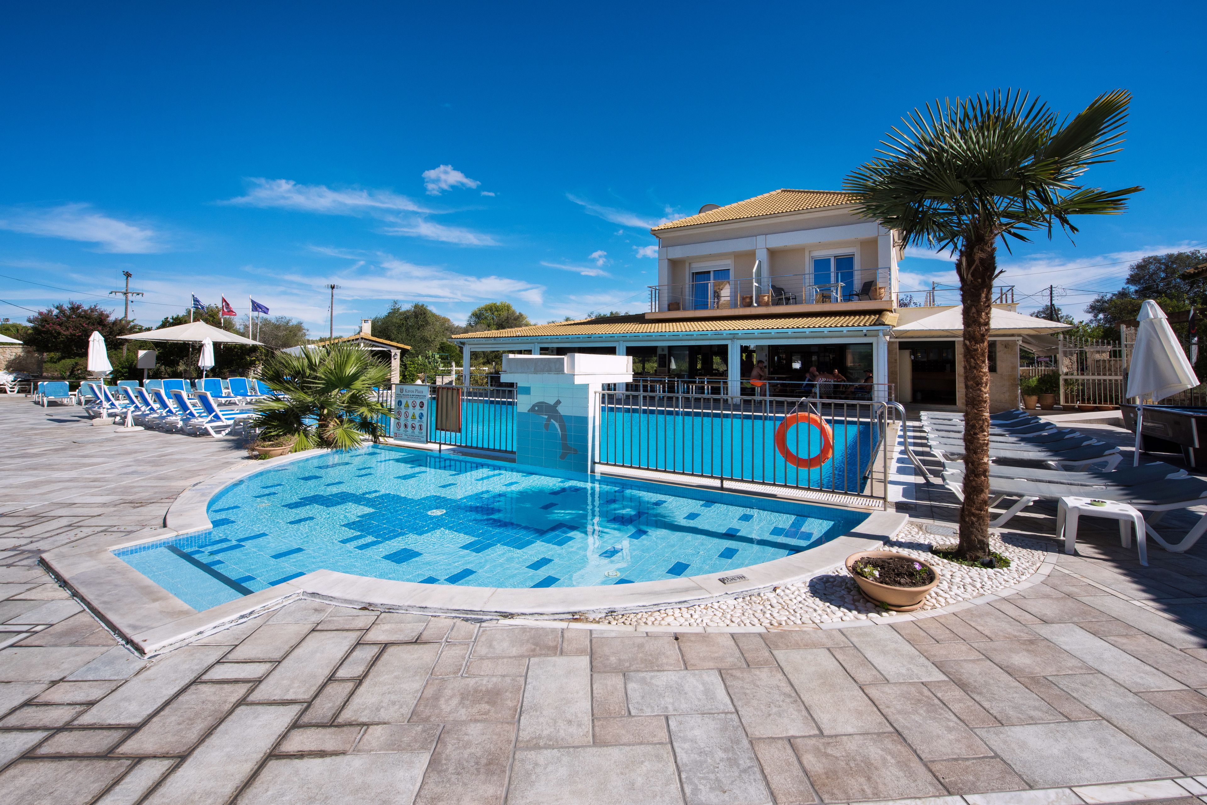 A swimming pool with lounge chairs and umbrellas, a palm tree, and a building with a balcony.
