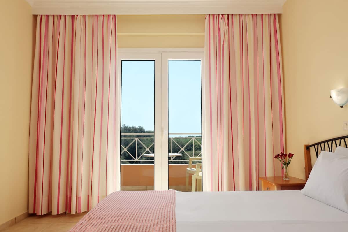 A hotel room with a bed, a nightstand with a vase of flowers, a window with curtains, and a balcony with a table and chairs.