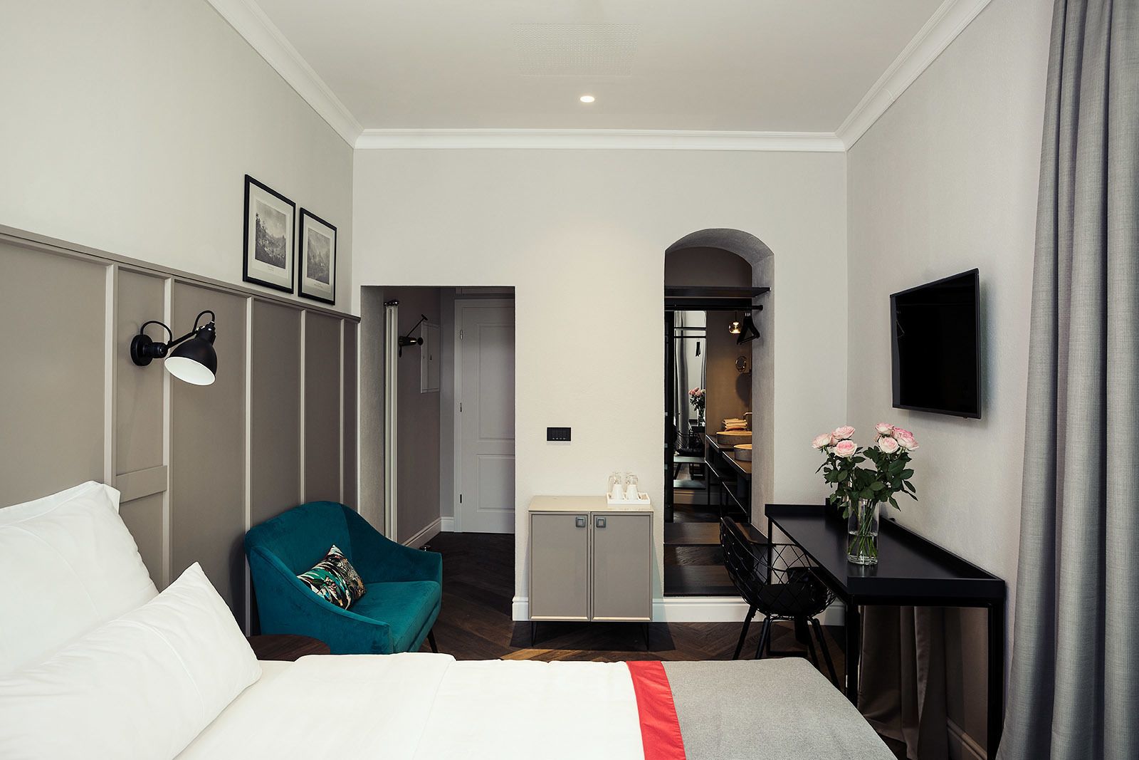 Une chambre d’hôtel avec un lit, un fauteuil turquoise, un bureau agrémenté d’un vase de fleurs et un téléviseur fixé au mur.