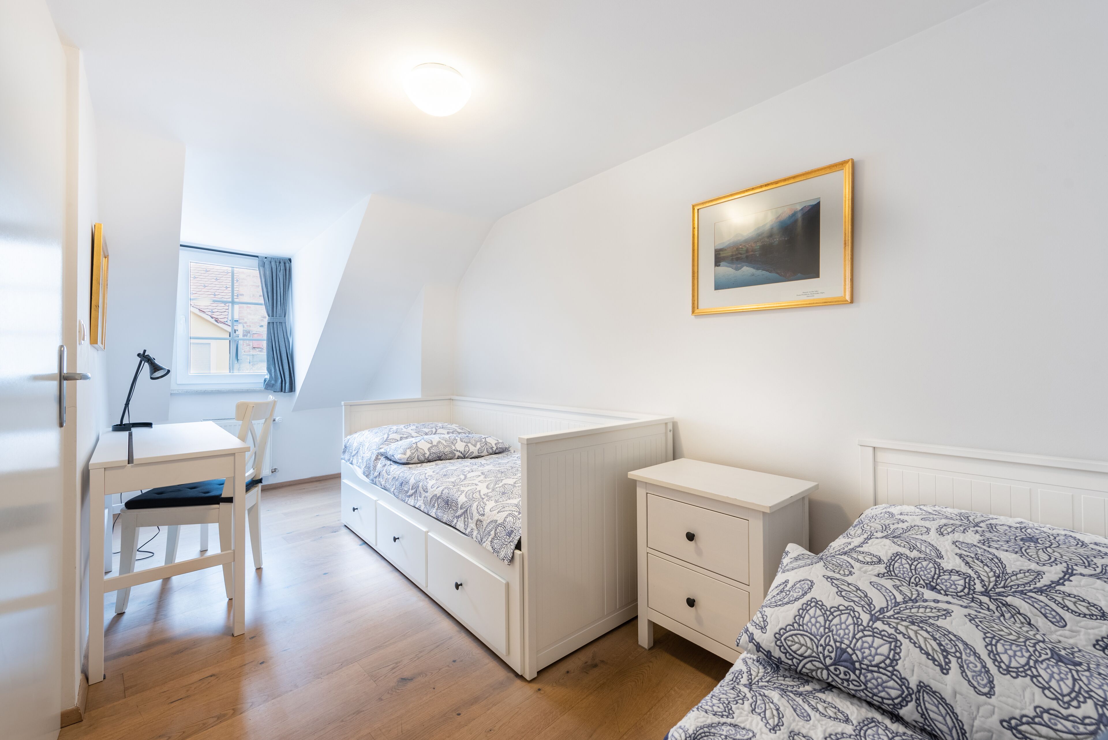 Een slaapkamer met een stapelbed, een bureau met stoel, een klein nachtkastje en een raam met gordijnen.