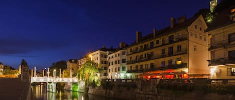 Blick auf eine historische Stadt an einem Flussufer, mit einer Brücke, Häusern mit Balkonen und einem ruhigen Fluss, der die Lichter reflektiert.