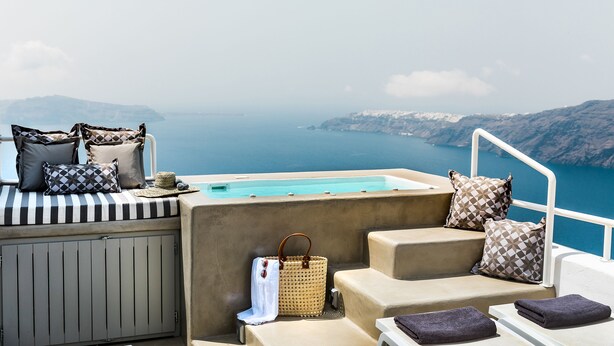 A sunlit outdoor seating area with a pool, beige and patterned cushions, and a woven basket. - 360 Caldera View Villa by Caldera Houses (Santorini)