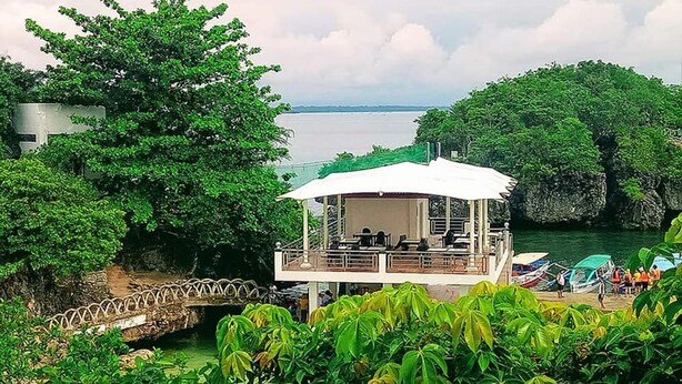 Uma estrutura estilo gazebo, de cor branca, com área de estar coberta, com vista para um corpo de água, cercada por vegetação exuberante e formações rochosas.