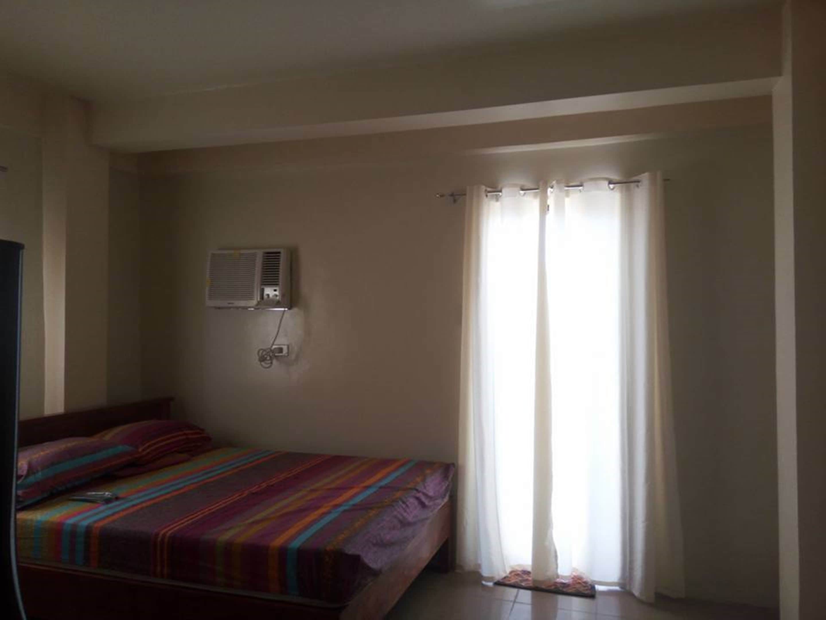 A bedroom with a bed, an air conditioning unit, a window with curtains, and a wall-mounted electrical outlet.
