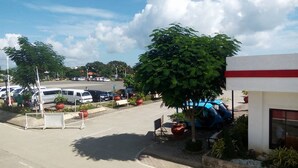 Um parque de estacionamento com vários carros, um edifício com uma faixa vermelha e uma árvore a dar sombra.