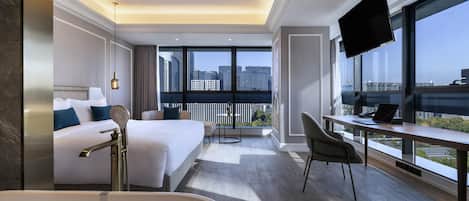 A modern hotel room with a large bed, a desk with a laptop, a TV, and a balcony with a city view.