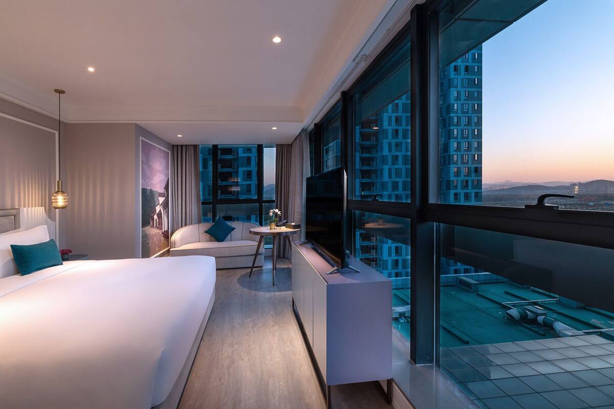 Habitación de hotel moderna con una cama grande, un sofá, una mesita y un televisor. La habitación tiene ventanas amplias que ofrecen vistas a la ciudad.