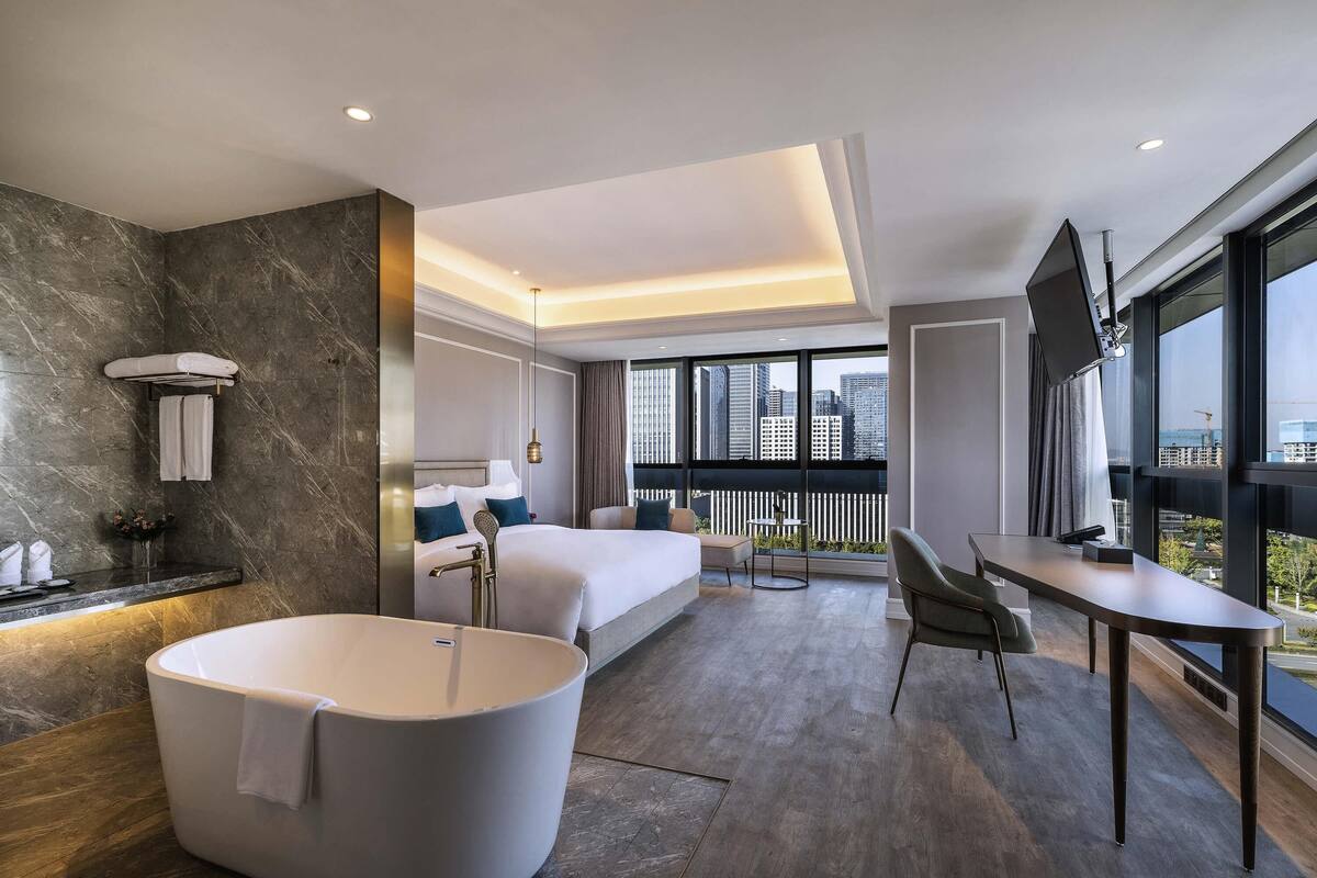 Una habitación de hotel moderna con una bañera grande, cama con vistas, escritorio y balcón con vistas a la ciudad.