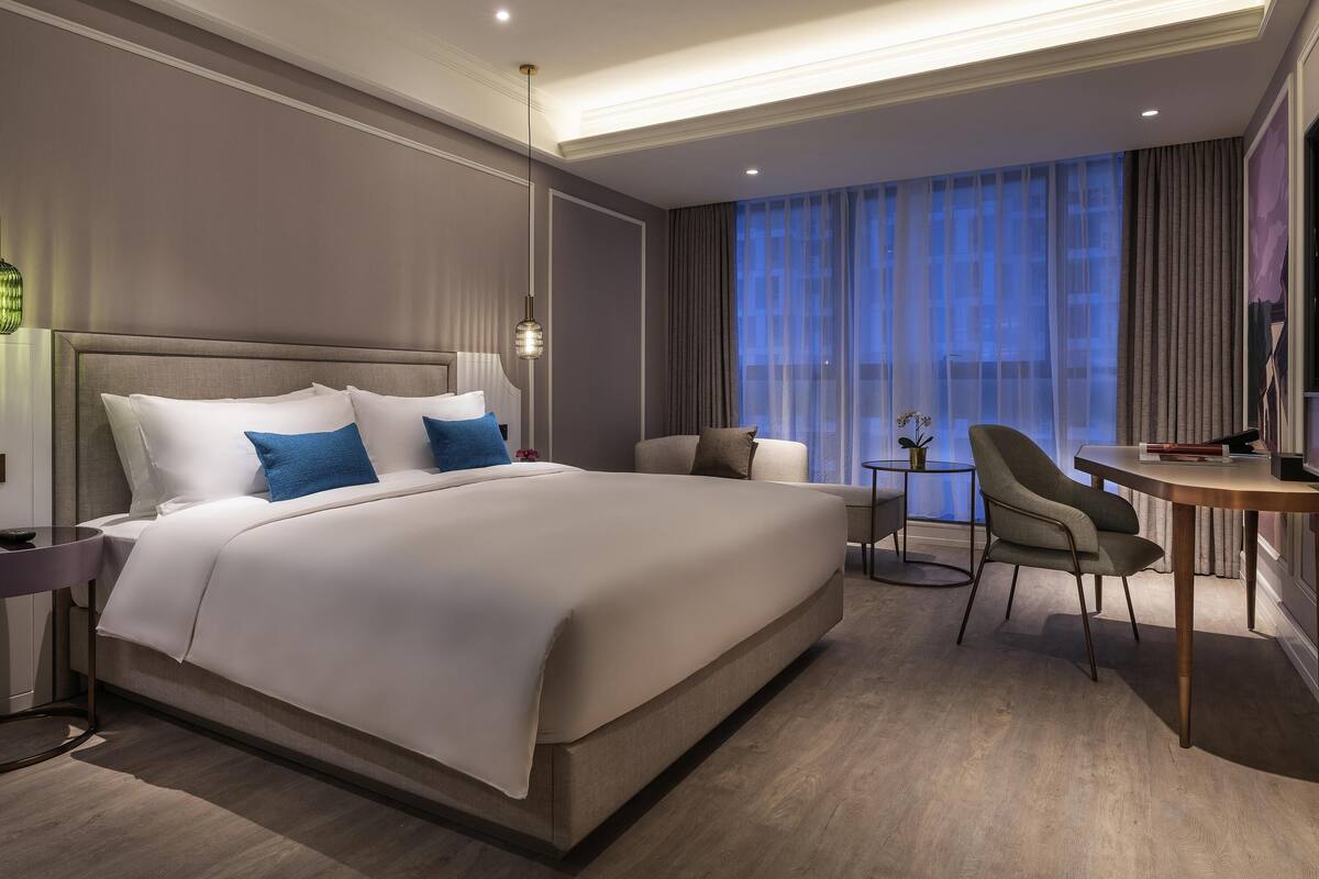 Una habitación de hotel moderna con una cama grande, un escritorio con silla, una mesita y un amplio ventanal con cortinas.