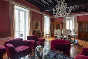 A room with a wooden ceiling, a chandelier, a dining table with chairs, and a red ottoman.