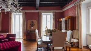 Een kamer met een eettafel, stoelen, een rode poef, een kroonluchter en een groot raam.