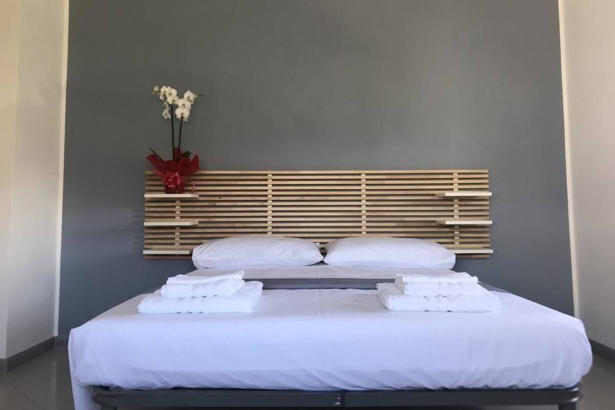 A hotel room with a bed, white linens, a wooden headboard, and a vase with white flowers.