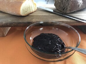 A glass bowl containing a dark substance, possibly a spread, on a wooden surface with a loaf of bread and a knife in the background. - Chiara's Home (Davoli)
