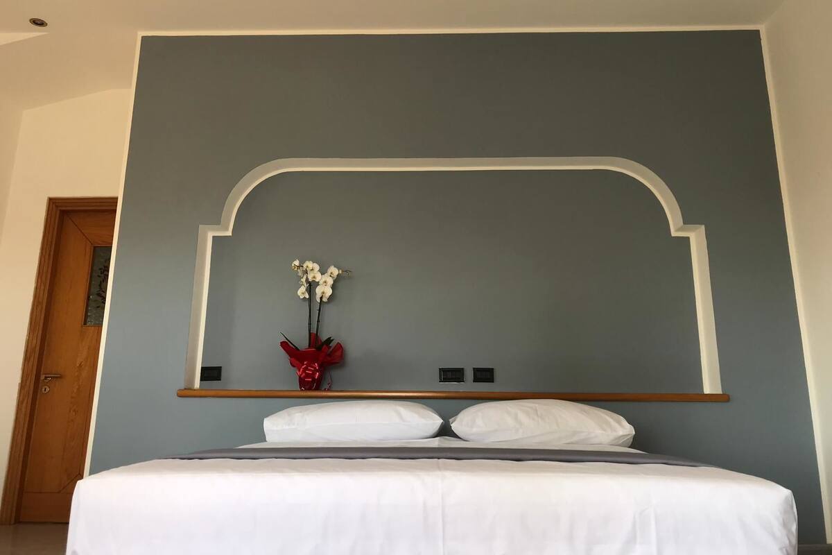 A hotel room with a large bed, a wooden headboard, and a wall-mounted shelf with a vase of flowers.