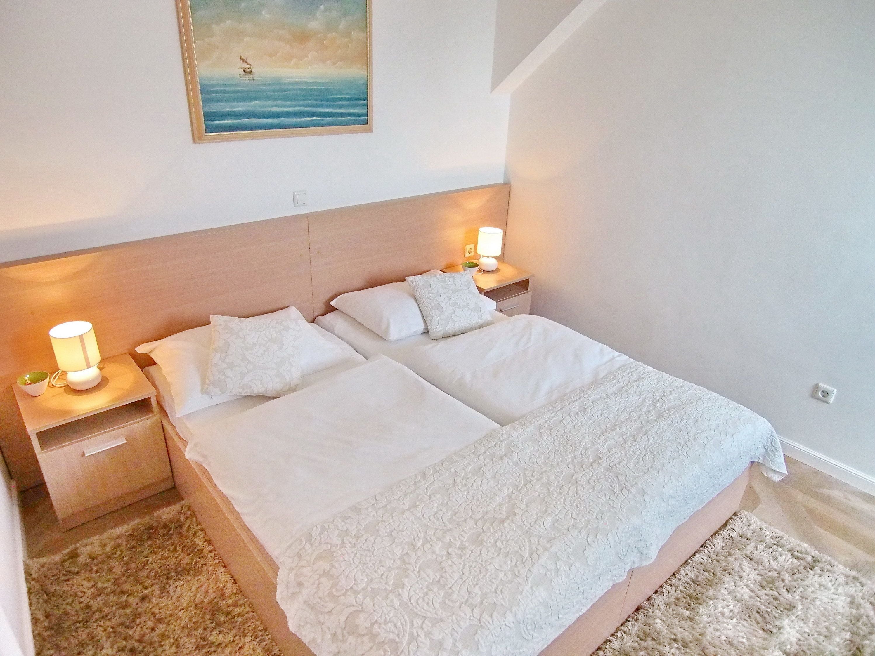 A double bed with white linens, two bedside tables with lamps, a small potted plant, and a painting of a sailboat on the wall.