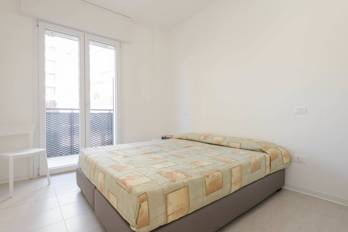 Quarto com cama, uma cadeira e porta de vidro corria para uma varanda.
