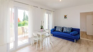 A bright living room with a white table and chairs, a blue sofa with patterned cushions, and large sliding glass doors leading to an outdoor area. - Color Apartments (Zupa dubrovacka)