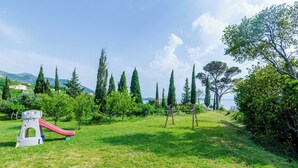 A grassy area with playground equipment, including a slide and swings, surrounded by trees and a clear sky. - Color Apartments (Zupa dubrovacka)