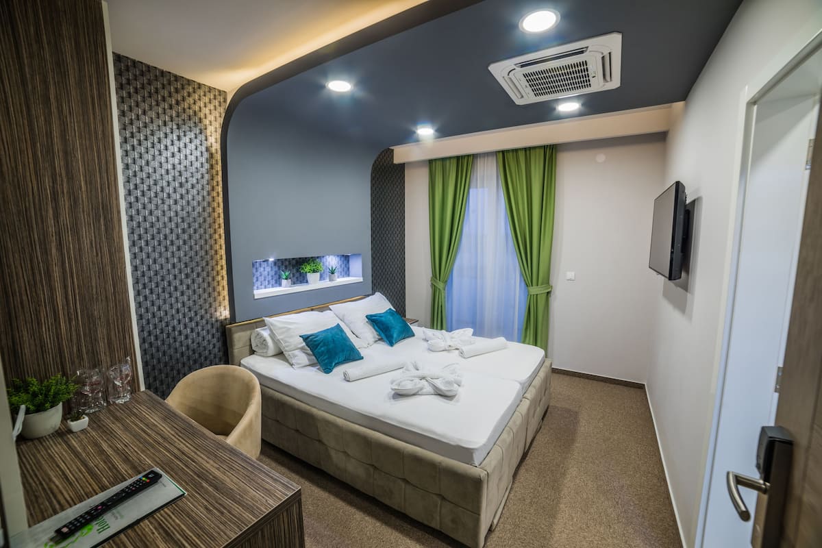 A modern hotel room with a large bed, a desk with a remote control, a TV, and a window with green curtains.