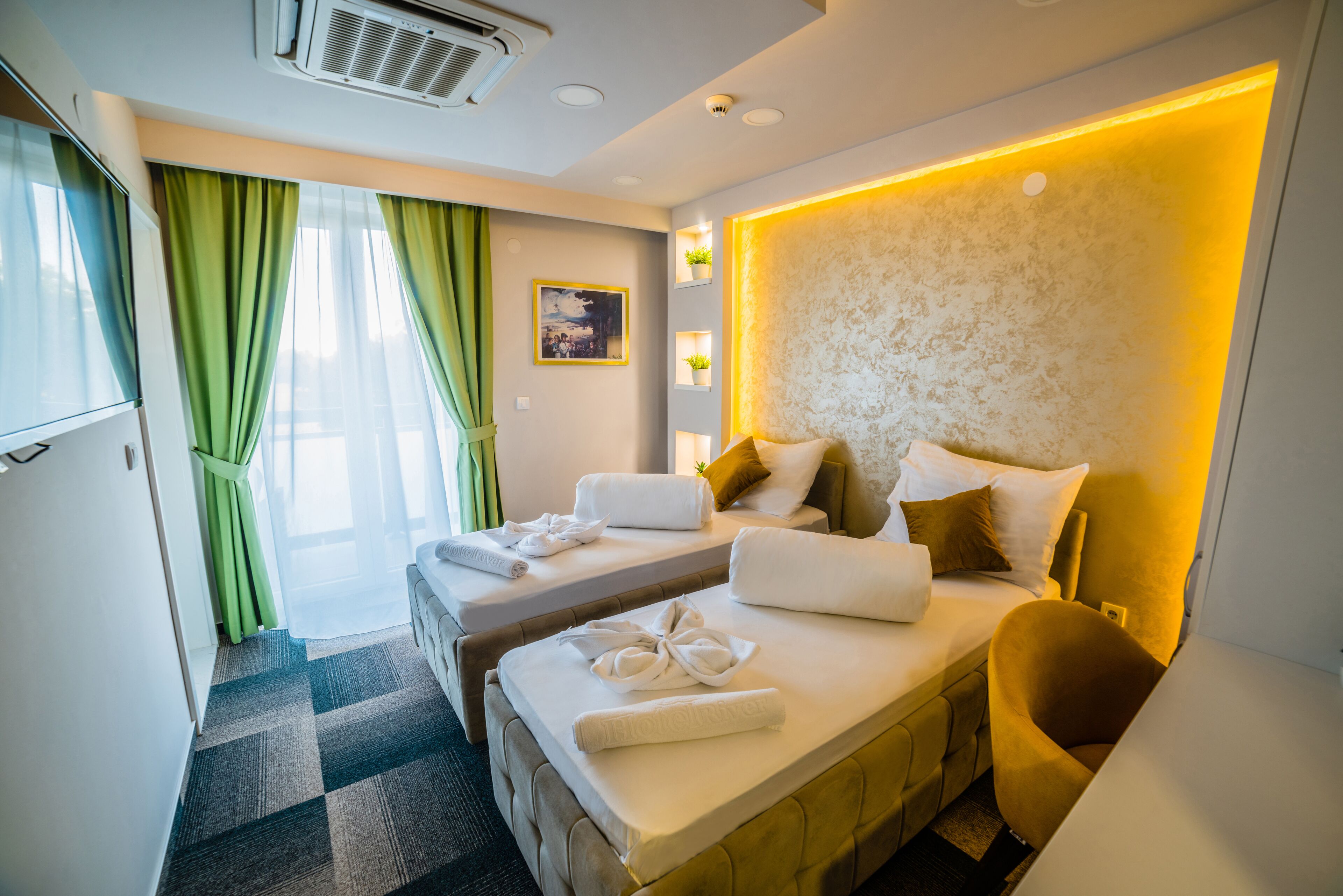 A hotel room with two beds, a sofa, a wall-mounted TV, a picture, and a small shelf with plants.