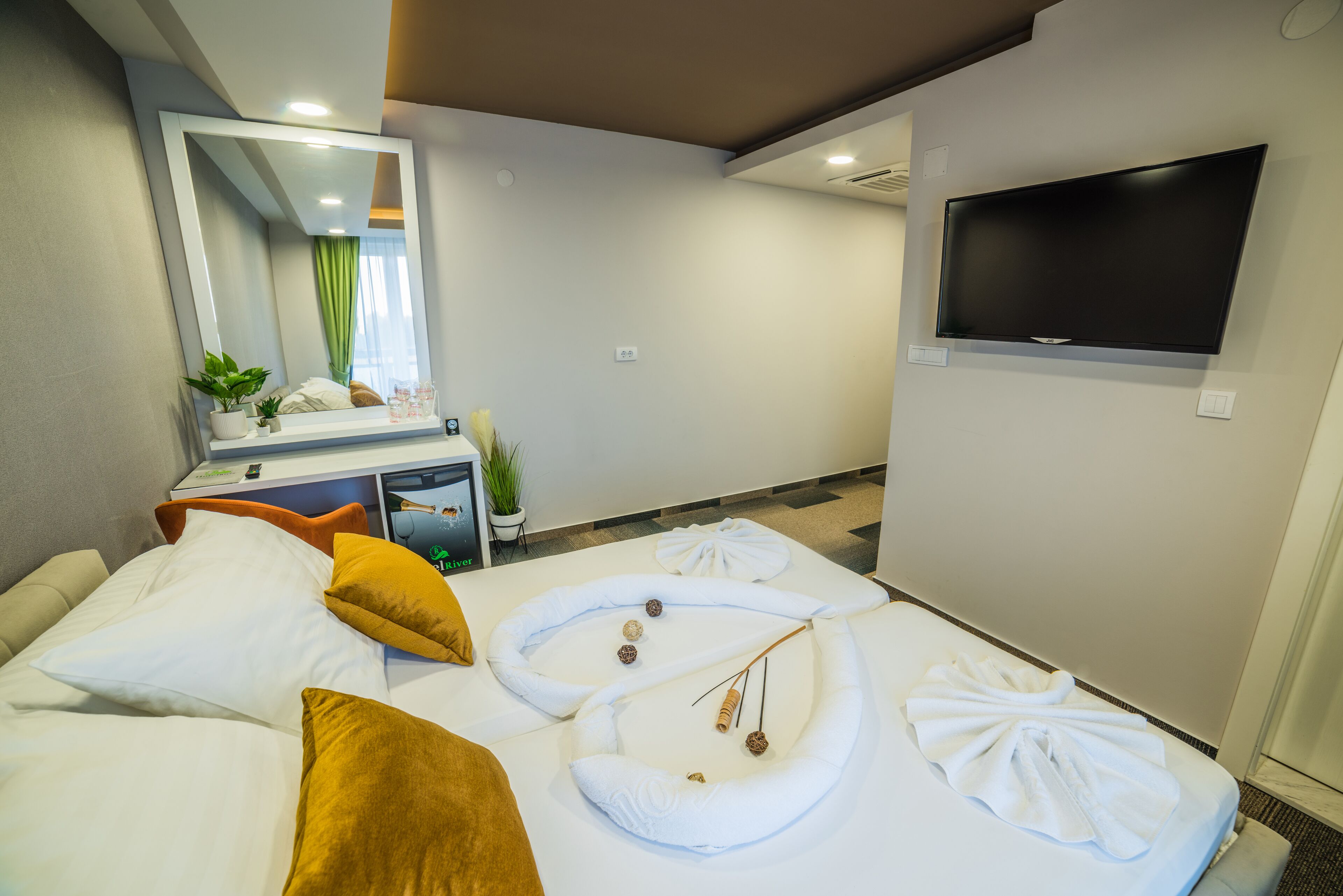 A hotel room with a large bed, a flat-screen TV mounted on the wall, a bedside table with a plant, and a mirror.