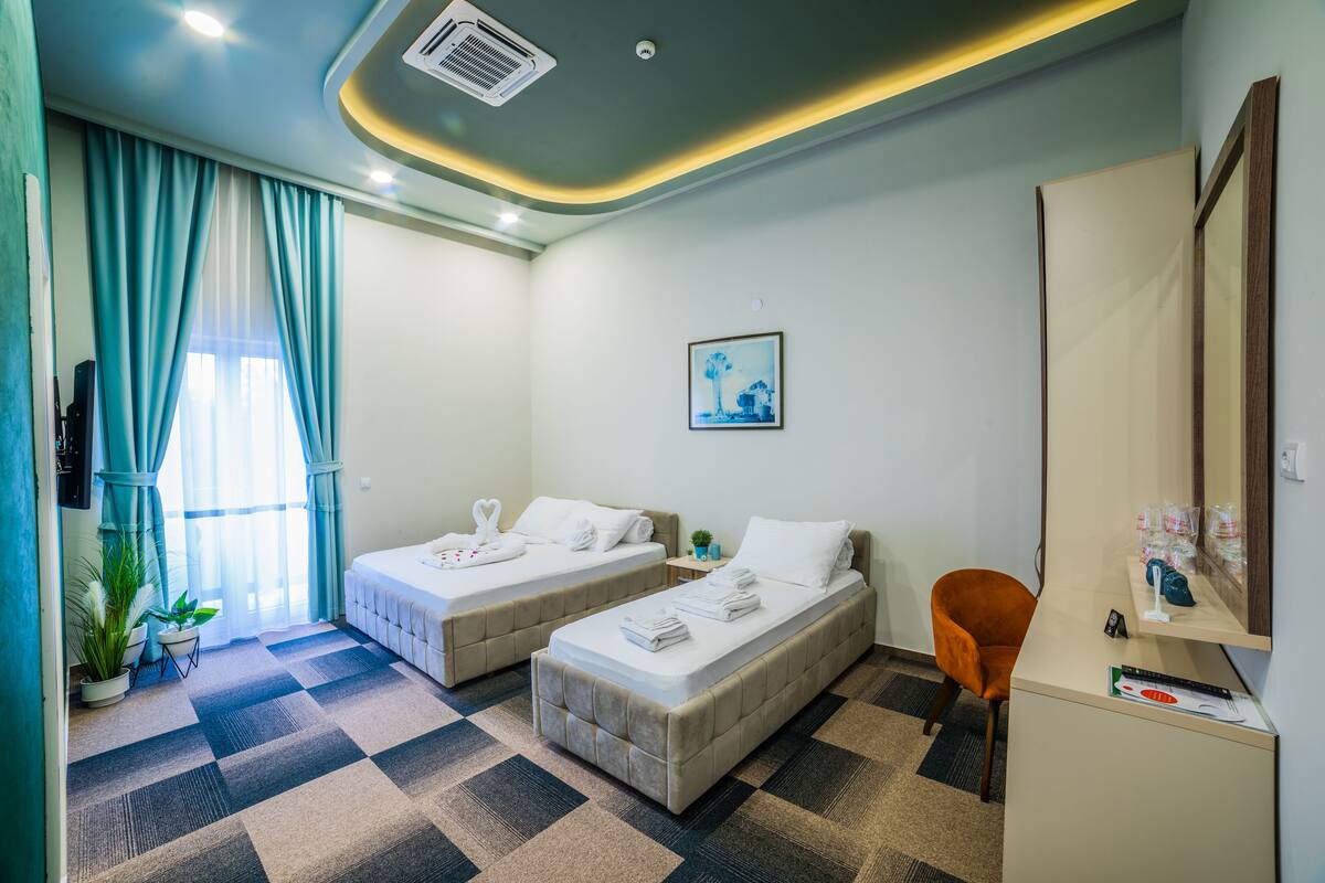 A hotel room with two beds, a desk, a chair, a potted plant, and a window with curtains.