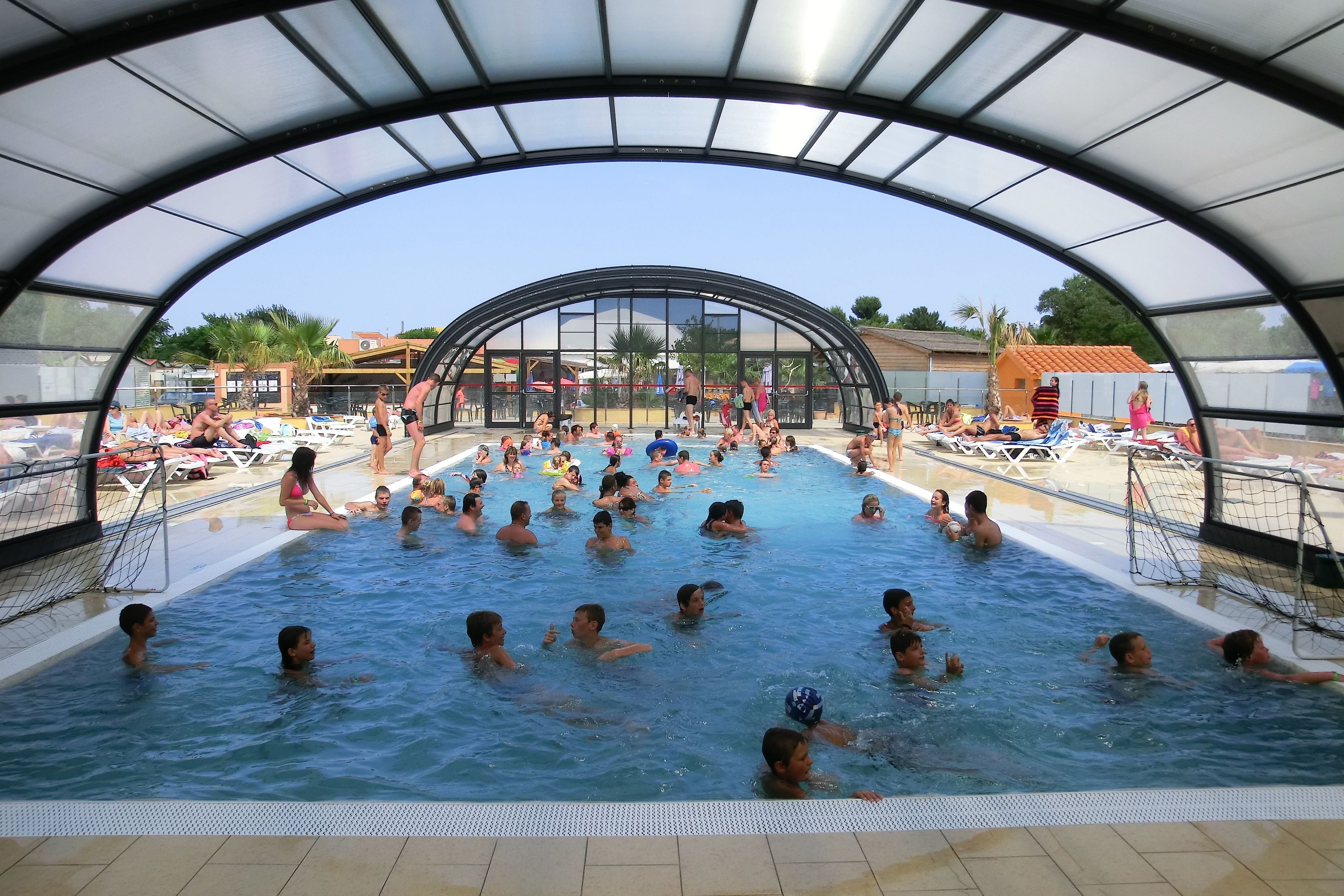 Ein Schwimmbad mit einer halbkreisförmigen Dachkonstruktion, umgeben von Liegestühlen und Sonnenschirmen.