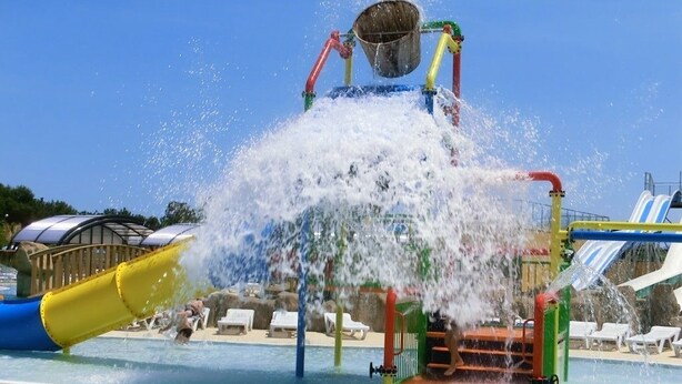 Un área de juegos acuáticos con un cubo grande y chorros de agua, rodeada por una piscina y toboganes.